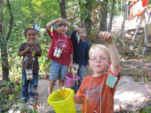 Group of four young children wear their shape bracelets on their wrist.  They are looking for nature object that match their shapes.  