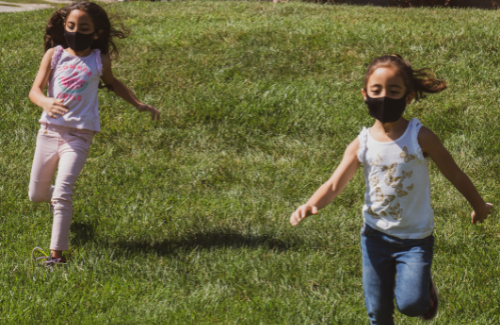 two young girls wearing masks running on grass outside