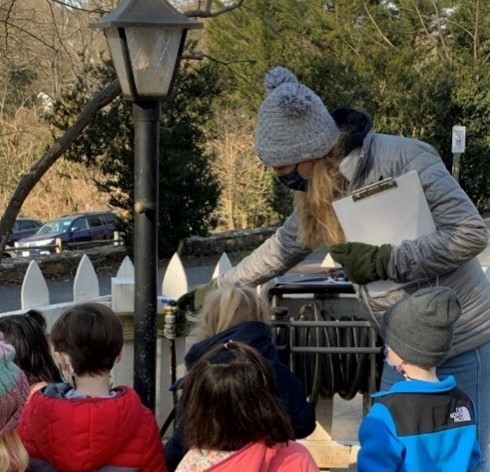 educator in a grey hat and jacket points to something outdoors while young children look on