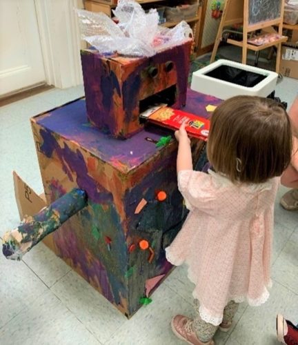 young girl feeds a handmade cardboard robot
