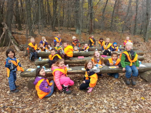 A group of diverse young students wearing orange safety jackets sit on homemade log benches in an outdoor classroom surrounded by leaves
