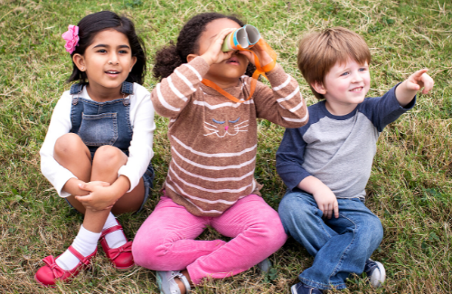 a young girl sitting on the grass looking into binoculars accompanied by another young girl and a young boy on each side