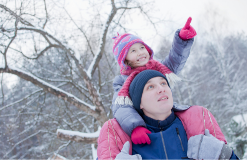 a young girl sitting on top of her father's shoulders as they walk near snow-covered trees wearing outdoor winter clothing pointing to the sky