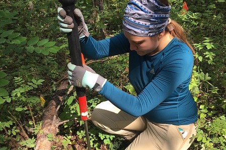 young woman kneels in a forest to sample soil using an auger