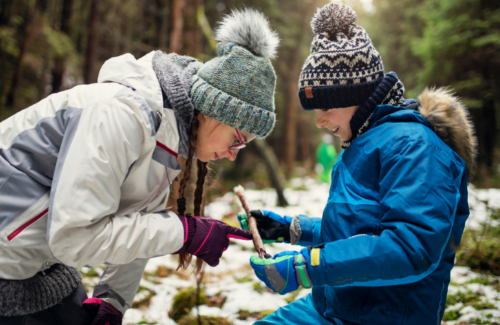 a woman and young boy dressed in winter clothing, including a hat, coat, gloves, and scarf observing a fallen tree branch in a snowy forest