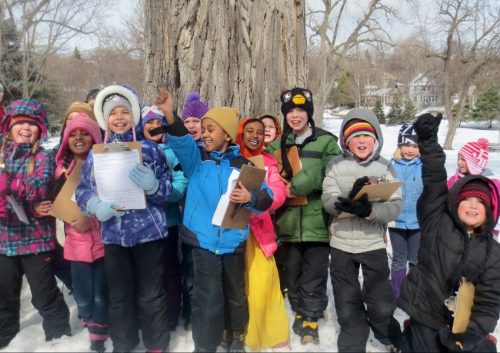 13 children wearing winter clothing standing in a line in front of a large tree trunk 
