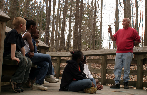 An educator surrounded by tall trees holding a leaf teaching 5 middle school students