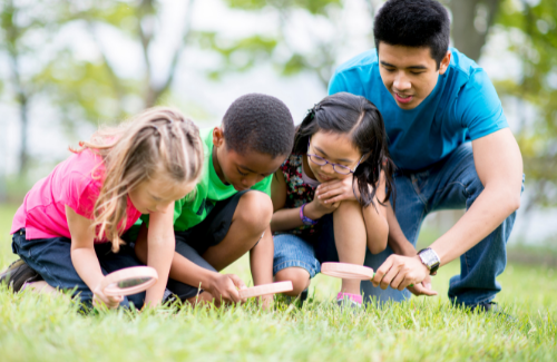 a teacher and three young students looking at the grass with magnifying glasses