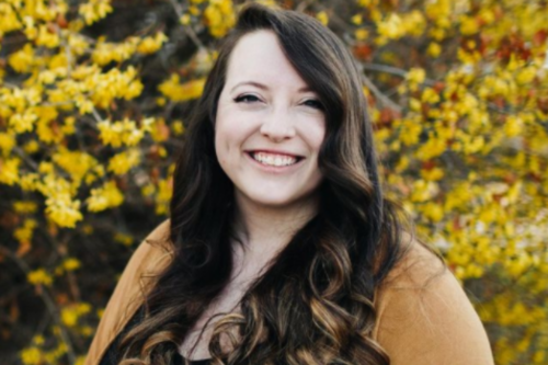 A headshot of Beth Hill smiling in front of a tree with yellow fall leaves