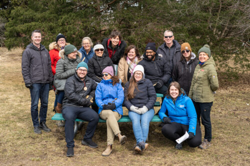 A group photo of 13 smiling new PLT facilitators outdoors in front of a forest